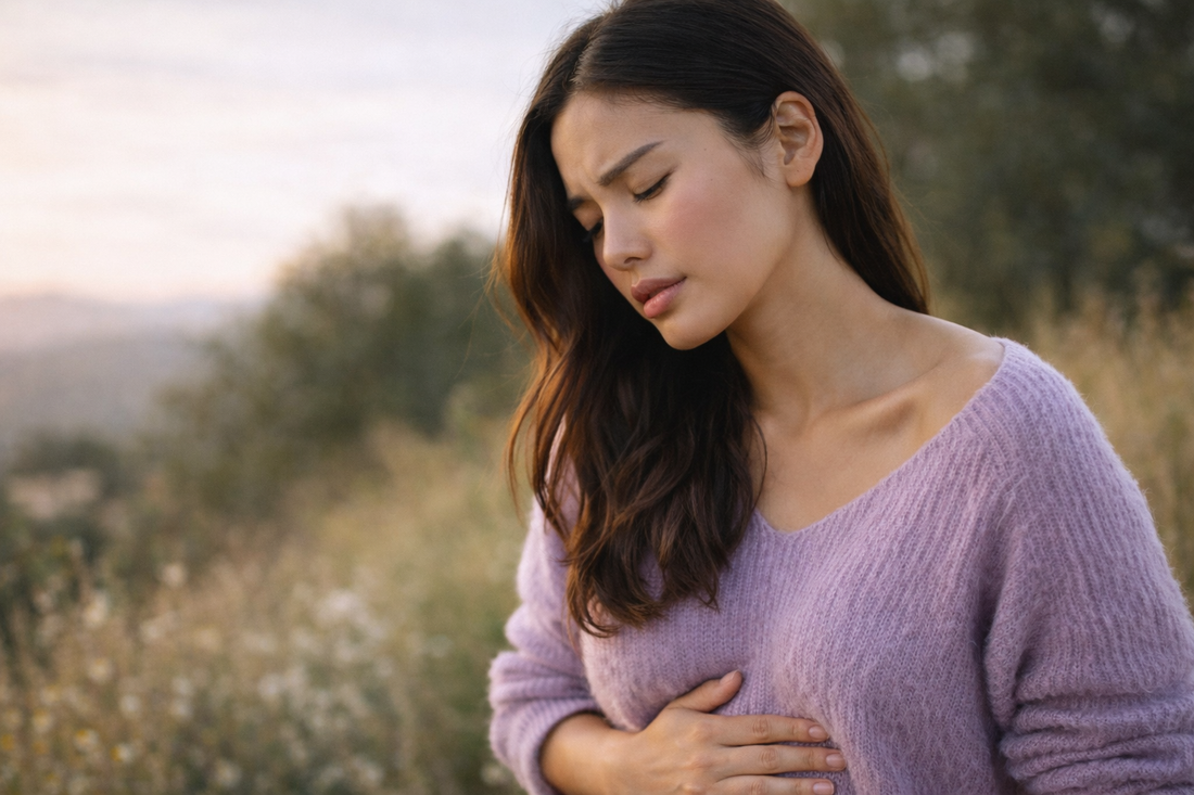 Femme debout dans un champ à la lumière dorée posant la main sur son ventre avec une expression légère d’inconfort dans une ambiance naturelle et apaisante